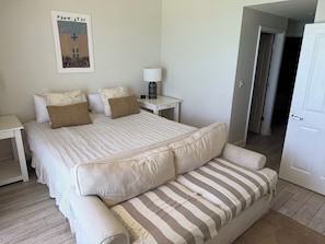 A neatly arranged bedroom with a large bed, a striped sofa, a bedside table with a lamp, and a door leading to another room.