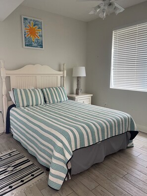 Quarto com cama, mesa de cabeceira, luminária, janela com persianas e um quadro na parede.
