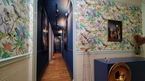 A hallway with wooden flooring, a blue wall with a bird and plant pattern, a framed picture, and a decorative vase.