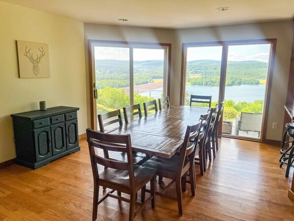 Uma sala de jantar com uma grande mesa de madeira e cadeiras, um armário de madeira escura e vista para um lago através de amplas janelas.