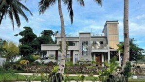 Une maison moderne à deux étages, dotée d’un toit plat, entourée d’un jardin bien entretenu avec des palmiers et diverses plantes.