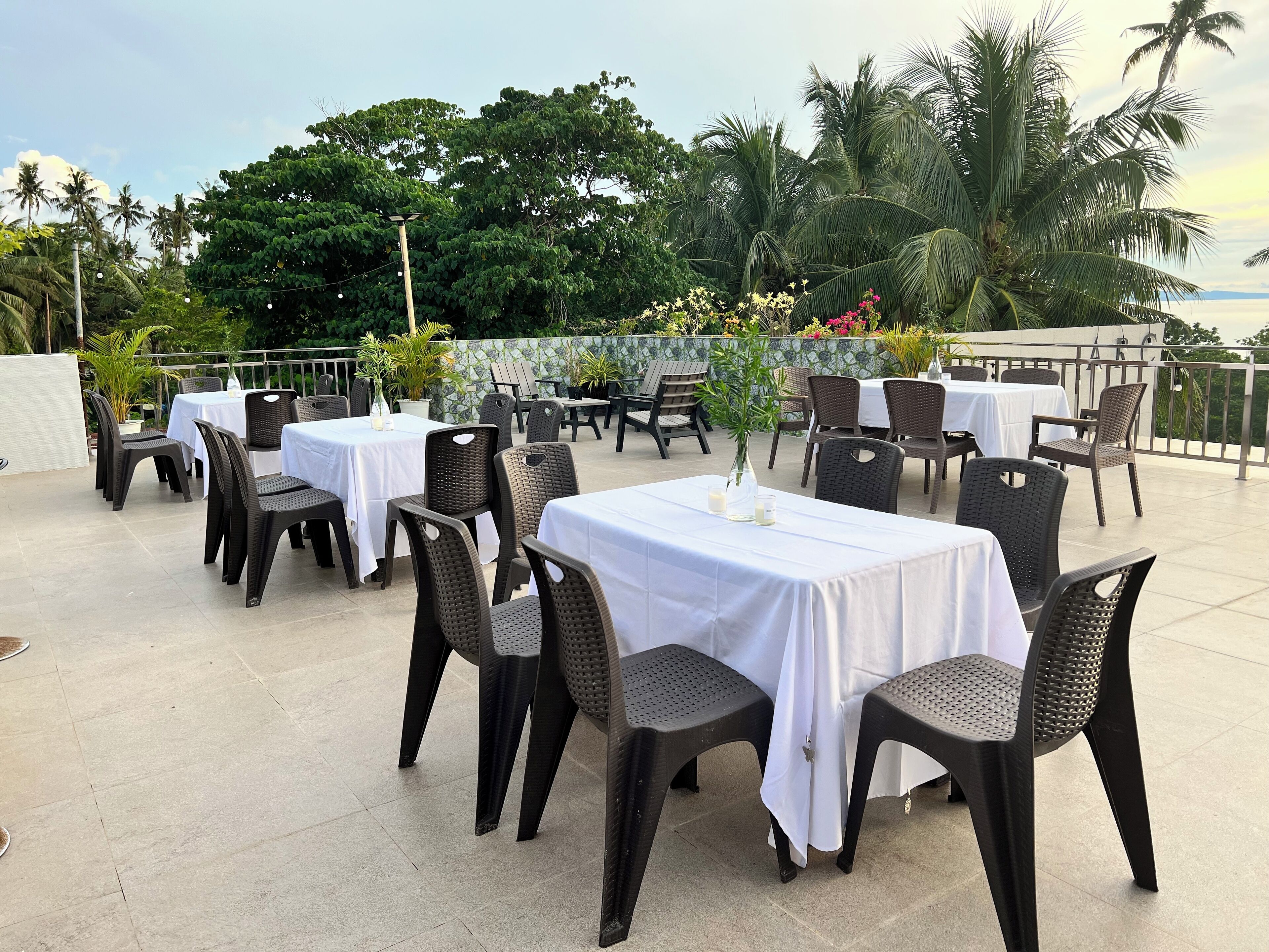 An outdoor dining area with tables and chairs, surrounded by lush greenery and palm trees.
