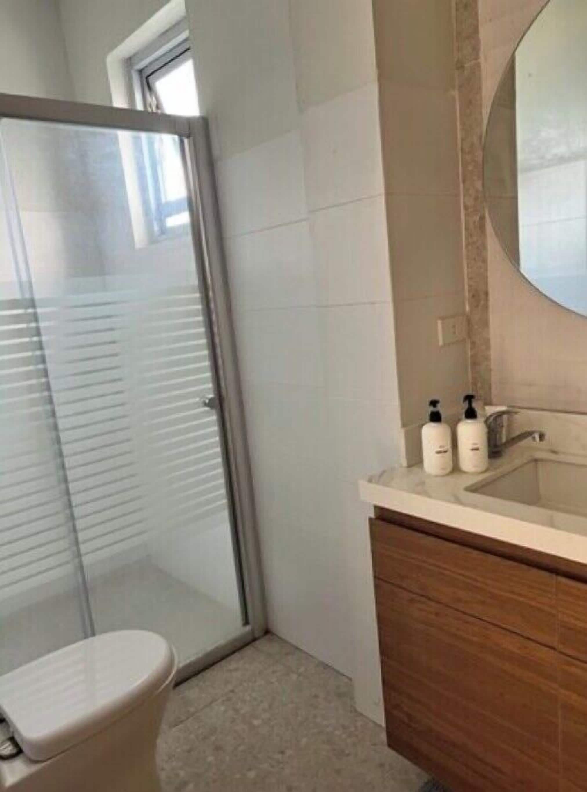 A bathroom with a toilet, a glass shower enclosure, a sink with toiletries, and a wooden vanity.