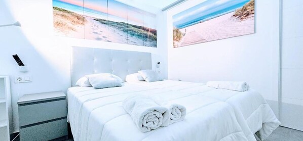 A hotel room with a large bed, white bedding, and two beach-themed wall art pieces.