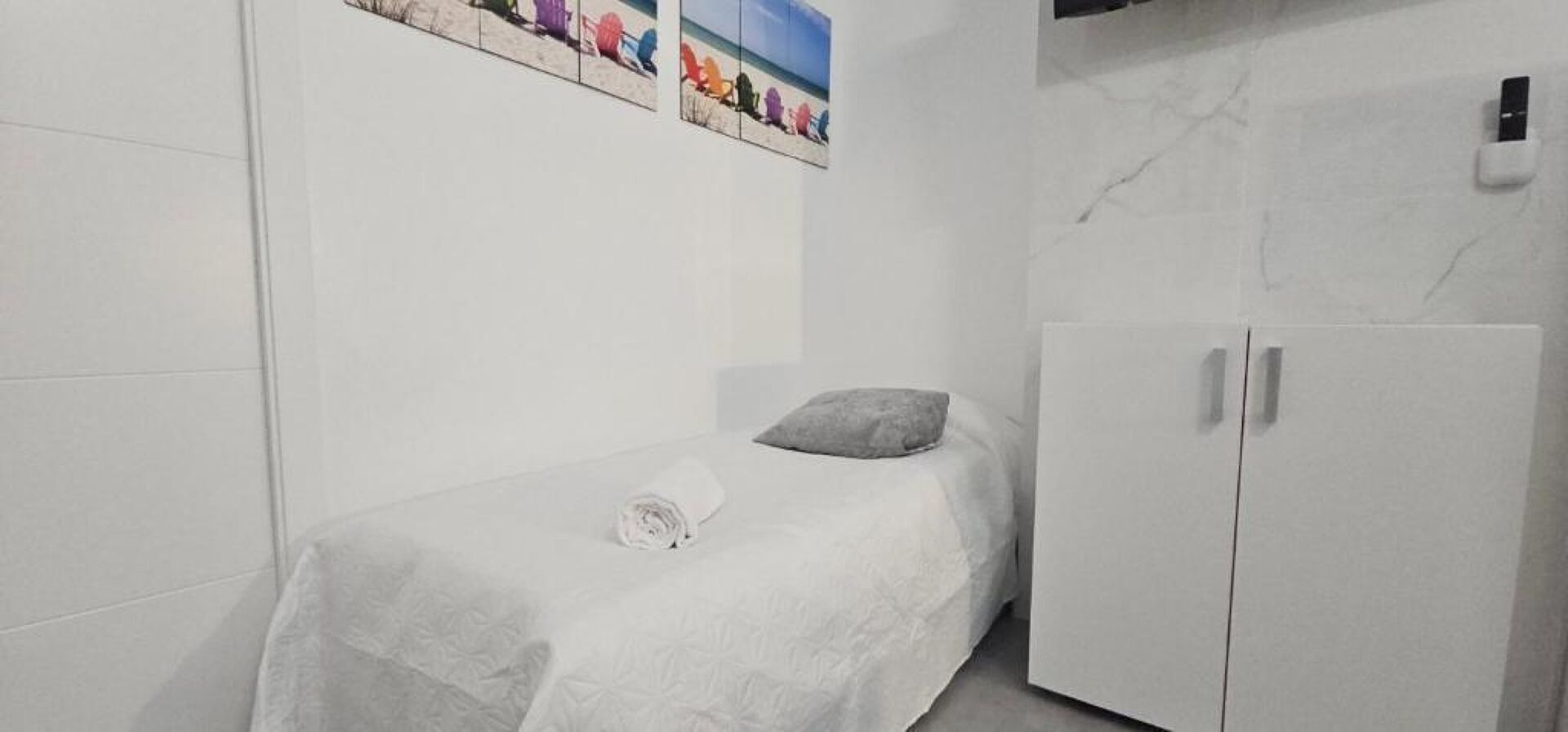 A small, neatly made bed with a pillow and a rolled towel, a white wardrobe, and a wall-mounted picture of beach chairs.