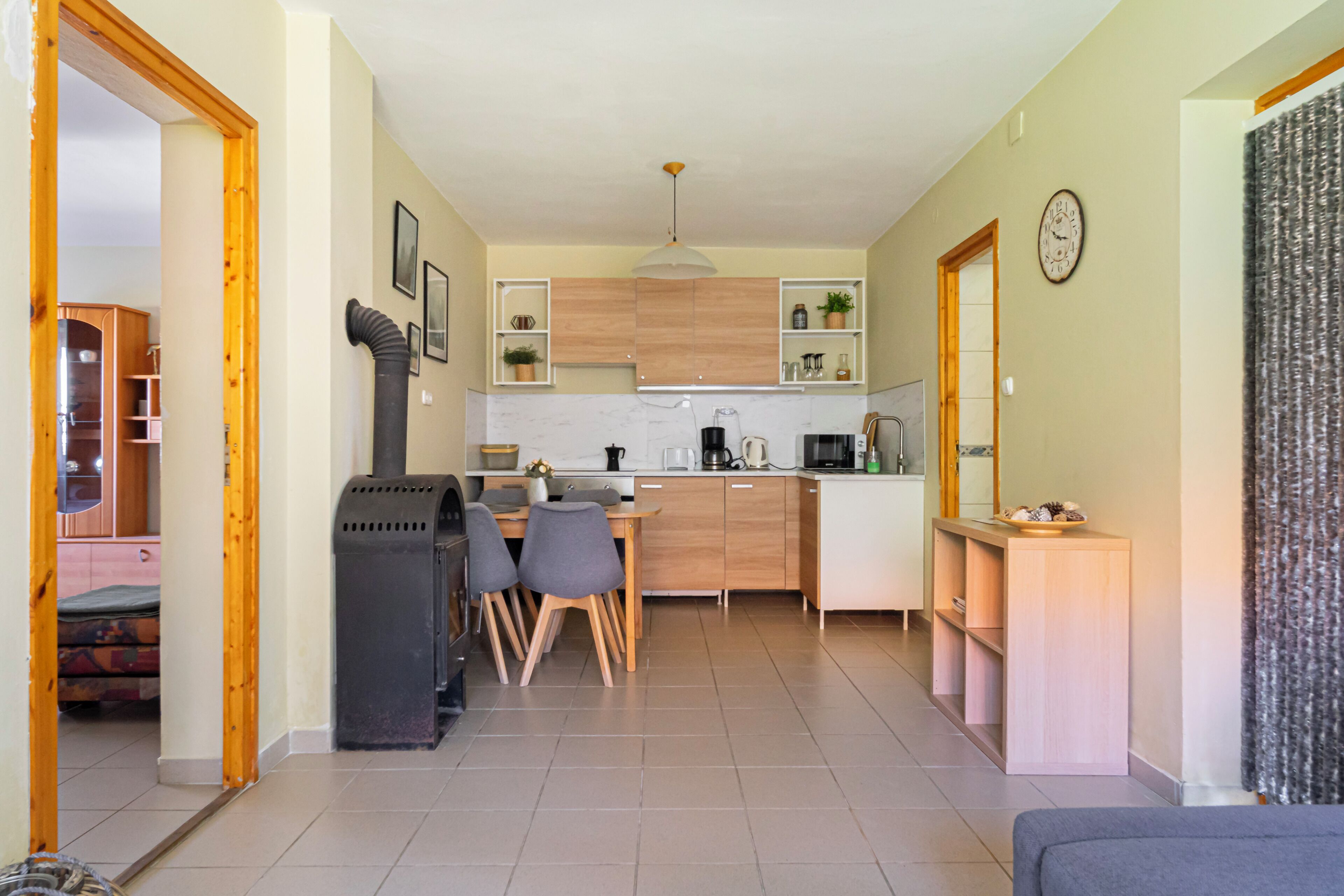 A compact kitchen with wooden cabinets, a dining area with chairs, a sofa, and a visible clock on the wall.