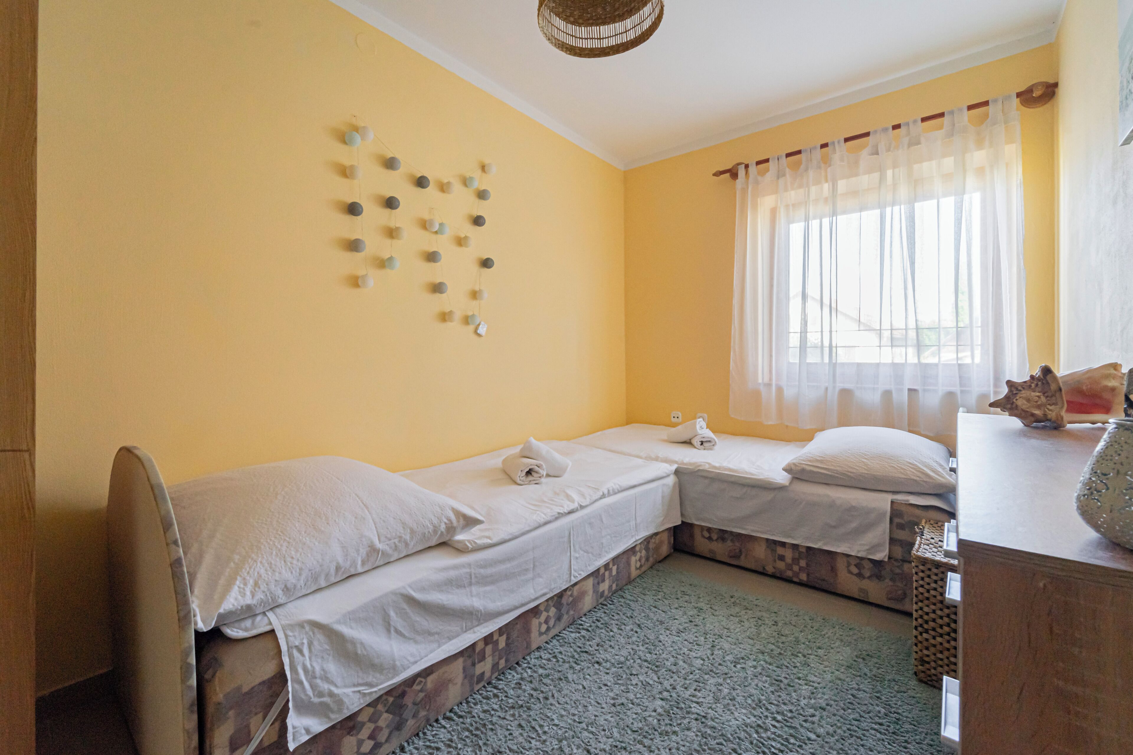 Ein Zimmer mit zwei Betten, einem Fenster mit transparenten Vorhängen, einer Wanddekoration und einem kleinen Tisch mit einem dekorativen Gegenstand.