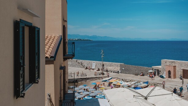 A coastal view with a stone wall, beach umbrellas, and a clear blue sea.