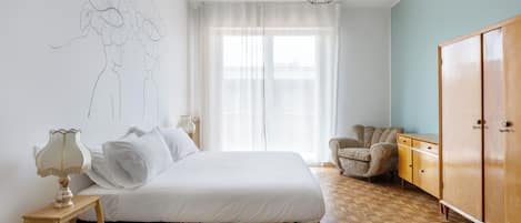 A bedroom with a bed, bedside table, lamp, chair, wardrobe, and a window with sheer curtains.