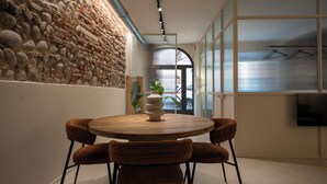 A modern dining area with a round wooden table and six brown upholstered chairs. A decorative vase is placed on the table. The room features a stone wall on one side and a large window with a view of the outside. - Suite Veronetta (Verona)