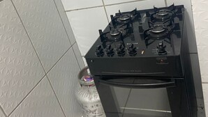 Una estufa negra con cuatro hornillas y un horno integrado, ubicada en un rincón de una habitación con paredes de azulejos blancos y una partición de vidrio.