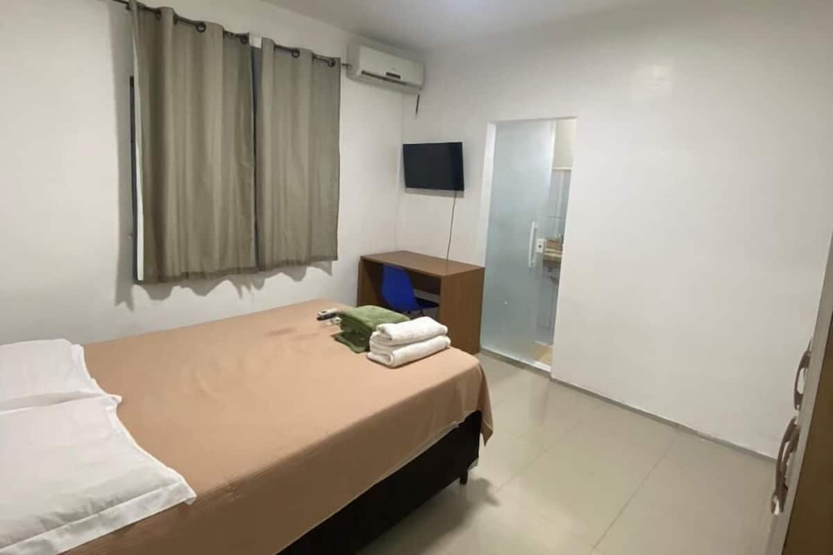 Ett hotellrum med en säng, en platt-TV som är monterad på väggen, en luftkonditioneringsanläggning, ett skrivbord med en stol och en skjutdörr i glas som leder till badrummet.