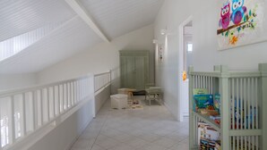 A children's playroom with a high ceiling, white walls, and a tiled floor. There is a bookshelf with toys and a door leading to another room. - Bel Embrun - Villa Iléenne (Les Trois-Ilets)