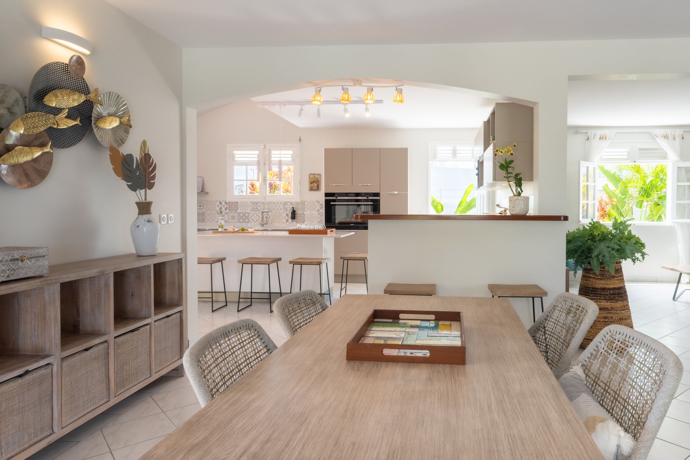 A modern kitchen with white cabinets, a central island, and a dining area with a long table and wicker chairs.