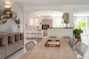 A modern kitchen with white cabinets, a central island, and a dining area with a long table and wicker chairs.