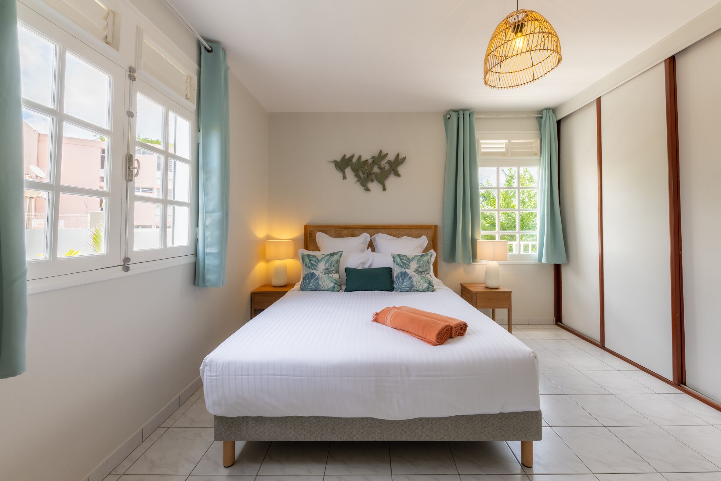 A neatly arranged bedroom with a large bed, two bedside tables with lamps, a hanging light fixture, and a view of the outside through the windows.