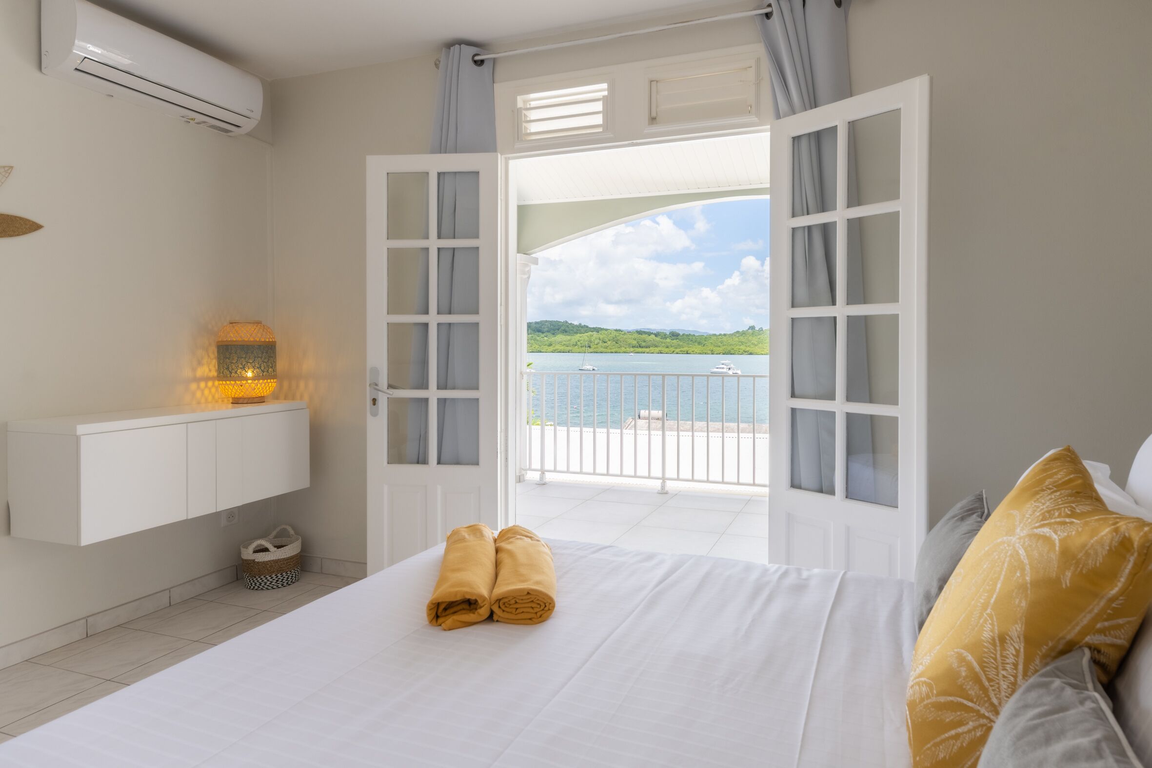 A bedroom with a bed, a nightstand, a wall-mounted air conditioner, and a view of the sea through open doors.