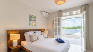 A bedroom with a large bed, bedside lamps, a view of the sea through an open door, and a tropical painting on the wall. - Bel Embrun - Villa Iléenne (Les Trois-Ilets)