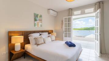 A bedroom with a large bed, bedside lamps, a view of the sea through an open door, and a tropical painting on the wall.