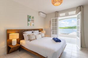 A bedroom with a large bed, bedside lamps, a view of the sea through an open door, and a tropical painting on the wall.