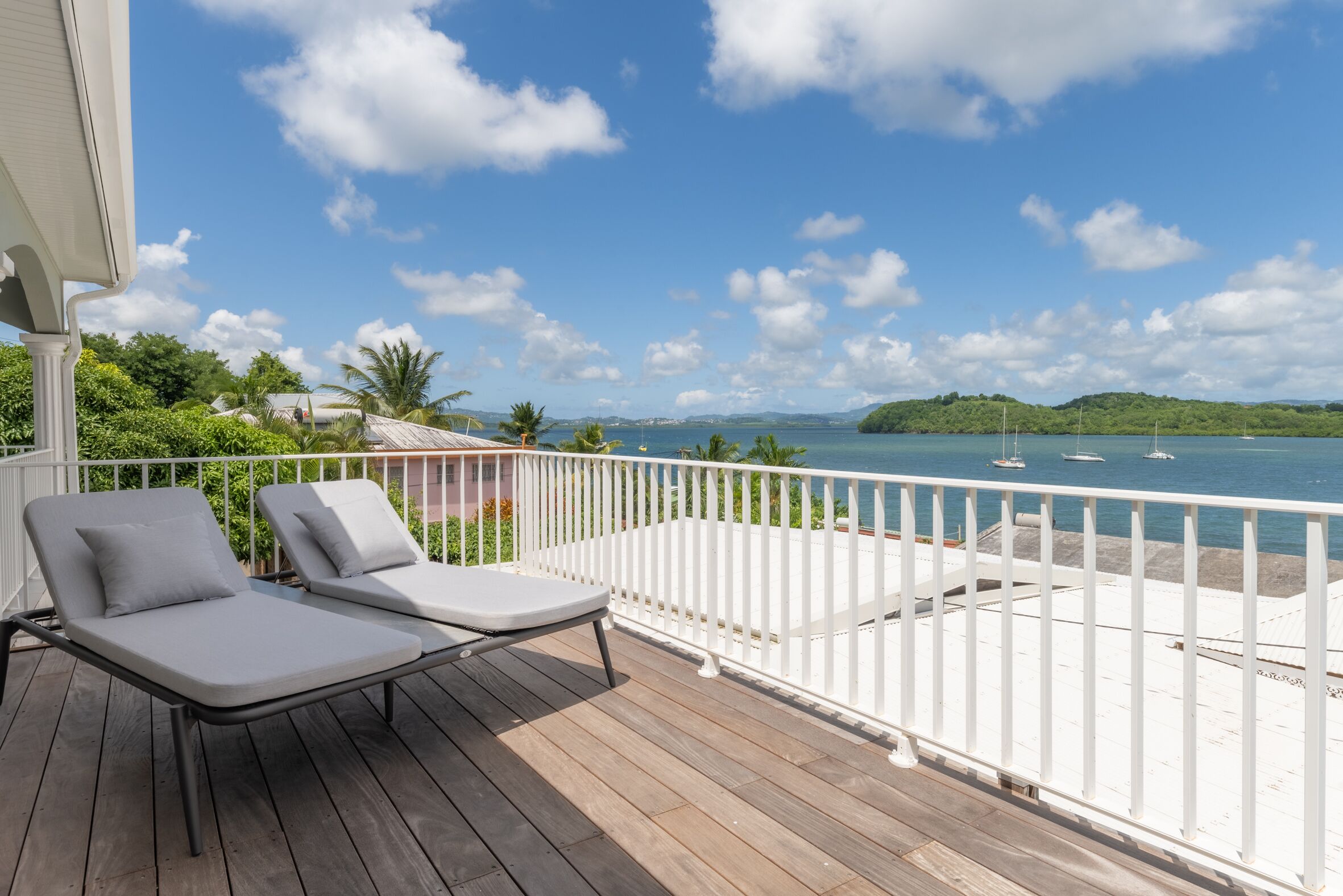 A wooden deck with two lounge chairs and a white railing overlooking a body of water with boats.