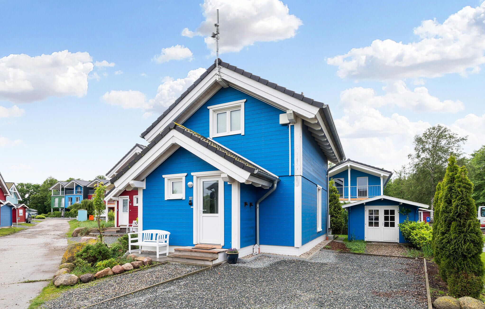 A blue house with a white door and windows, a small porch with a bench, and a gravel driveway.