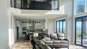 A spacious living area with a high ceiling, a balcony with a railing, and a kitchen in the background.
