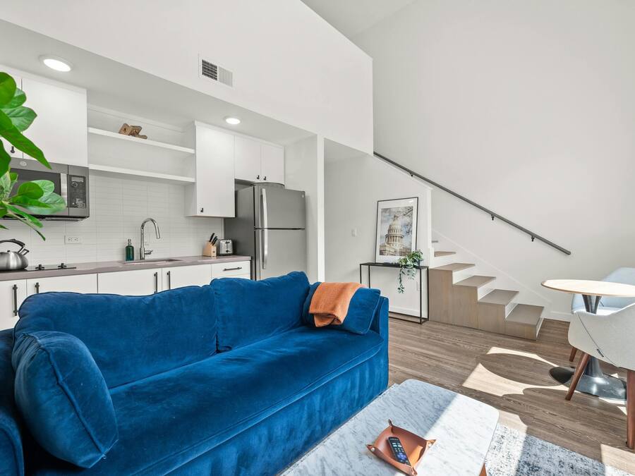 A modern living space with a blue sofa, a kitchen with white cabinets, a staircase, and a dining area with a white table and chairs.