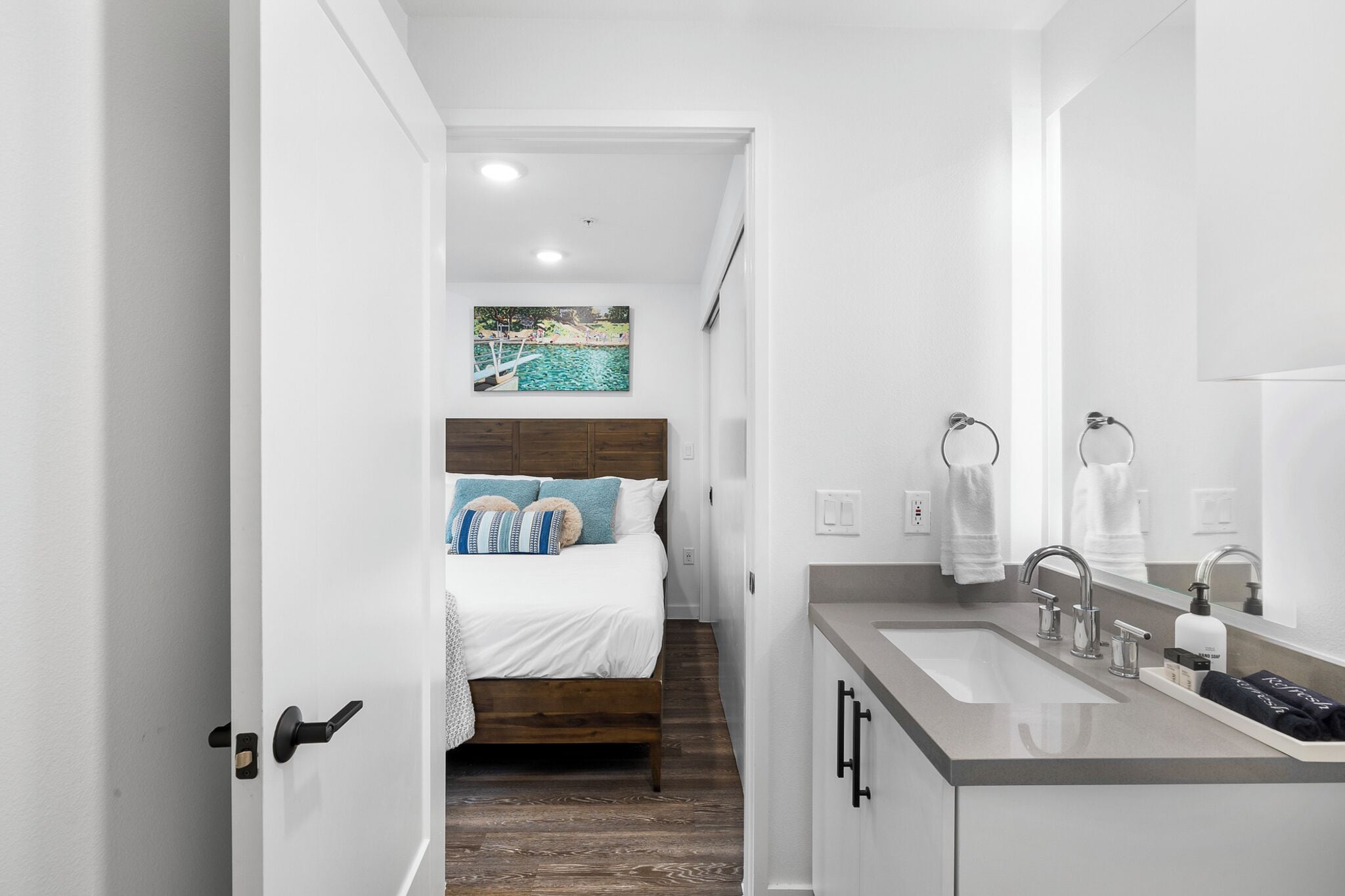 A narrow hallway leading to a bedroom with a bed, a bathroom with a sink and mirror, and a painting of a beach scene on the wall.