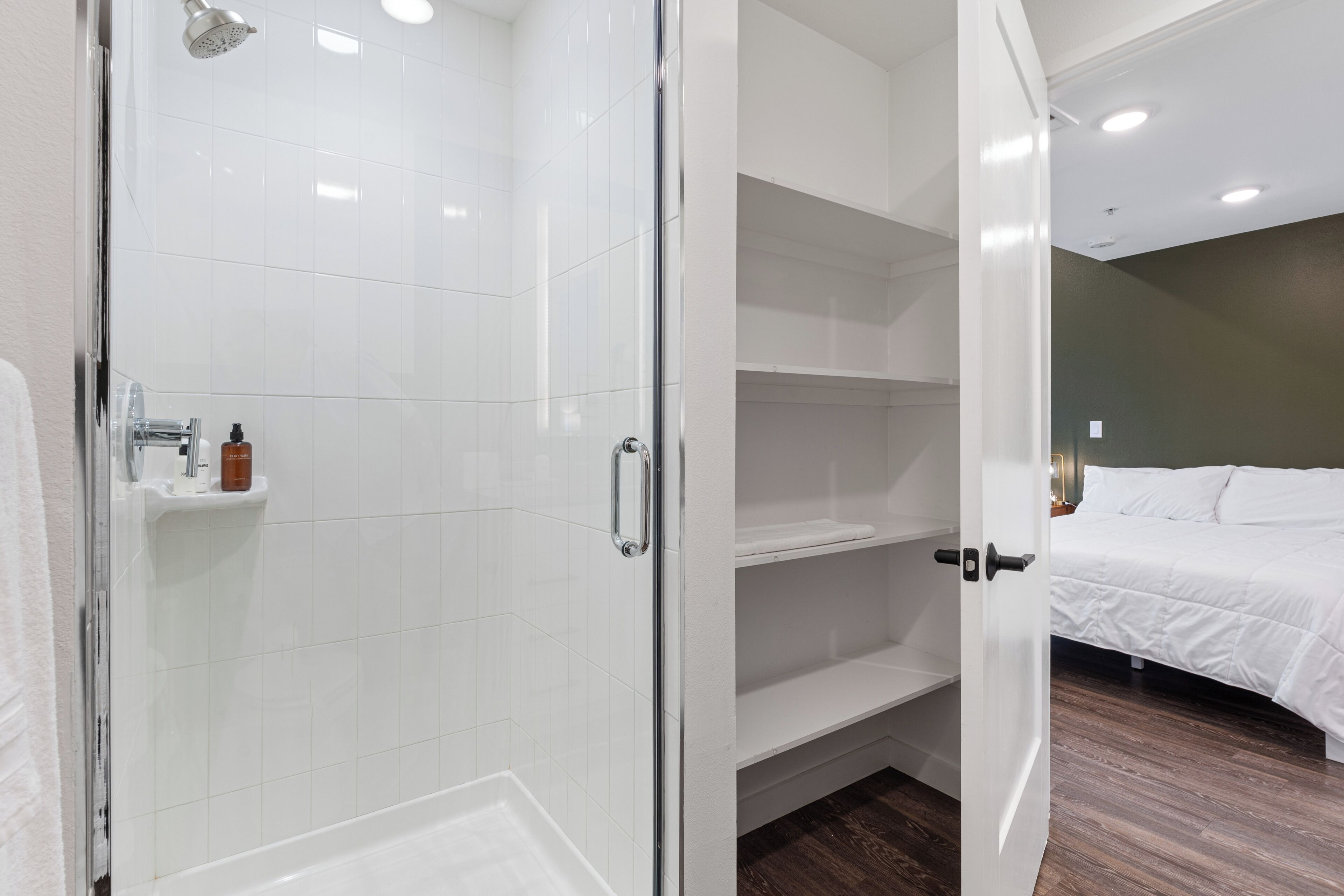 Une salle de bain moderne avec une douche en verre, un lavabo blanc et une étagère contenant des articles de toilette. À côté se trouve une chambre avec un lit et un plancher en bois.