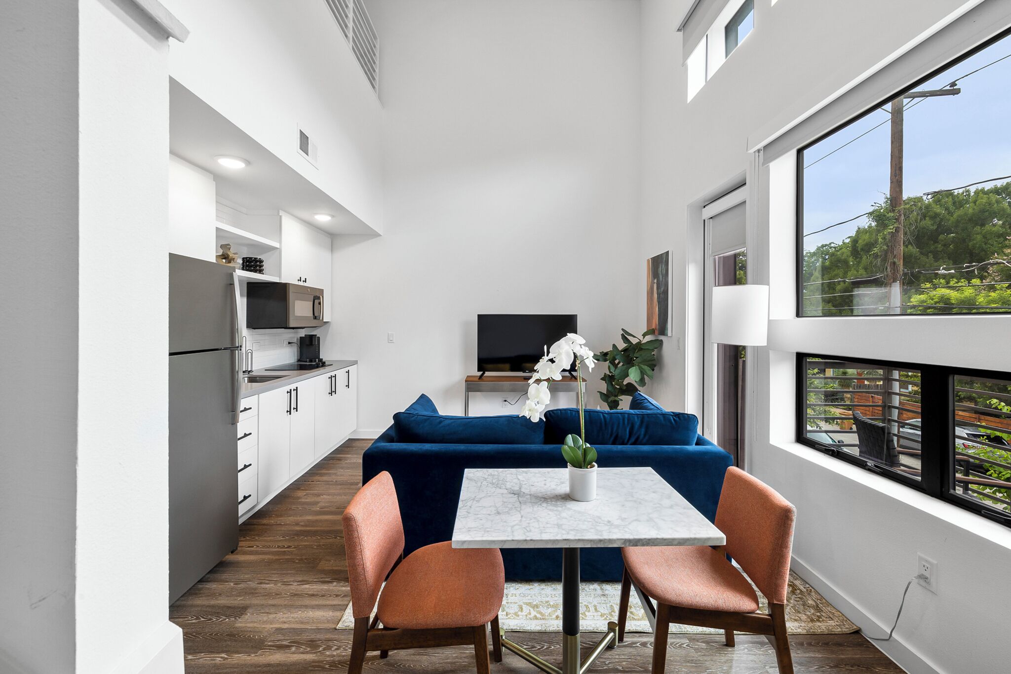 Una cocina moderna con armarios blancos, un microondas y un televisor. Hay un sofá azul y una mesa con sillas en la zona de estar.