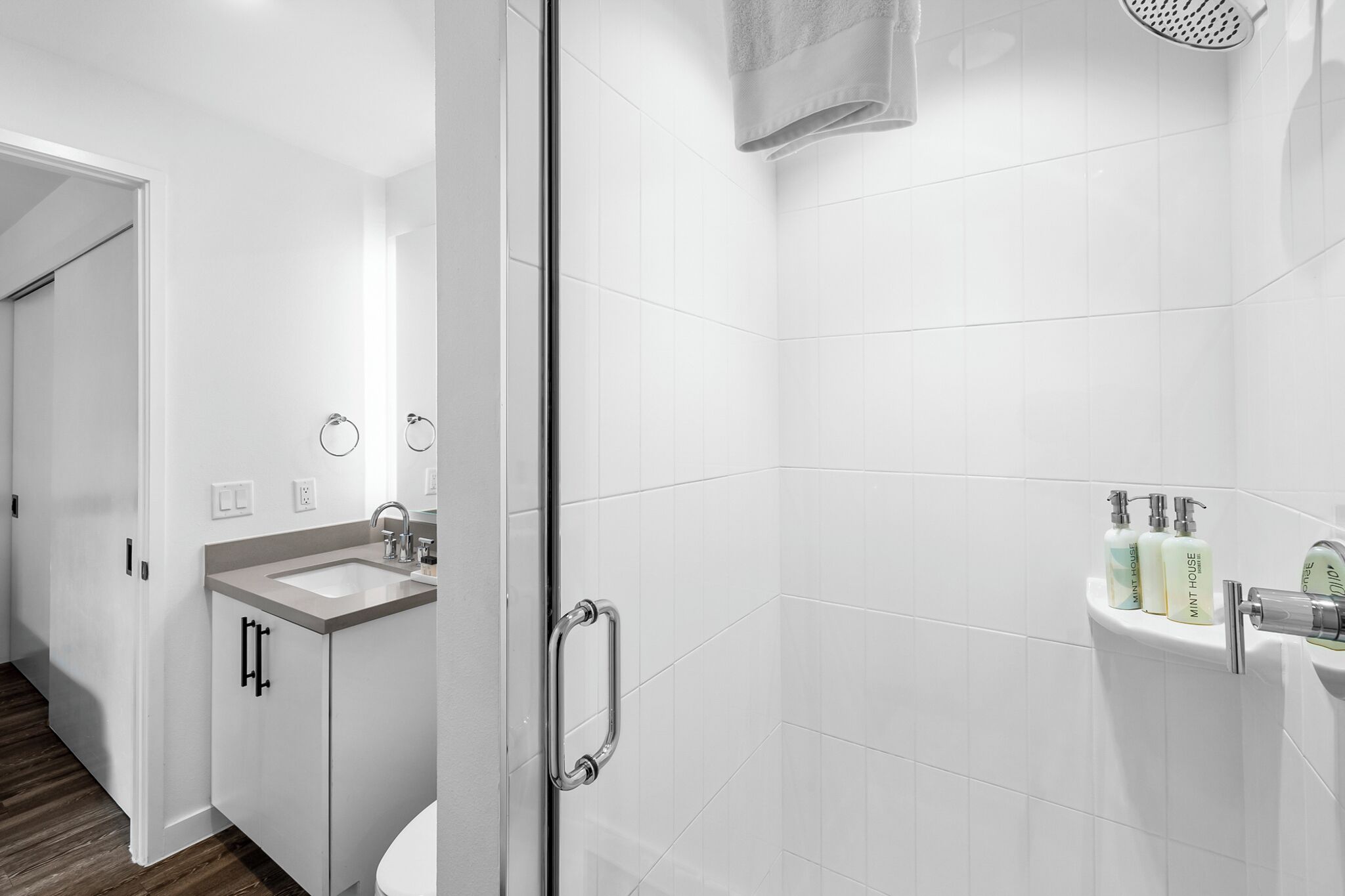 Une salle de bain moderne avec une douche à parois de verre, un lavabo et des toilettes. On peut voir des articles de toilette accrochés au mur et une serviette suspendue à la porte de la douche.