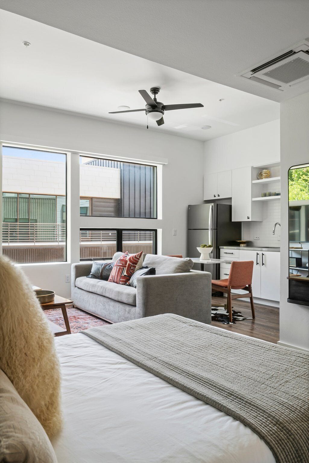 A modern, well-lit room with a grey sofa, a dining table, and a kitchen area with white cabinets and a refrigerator.