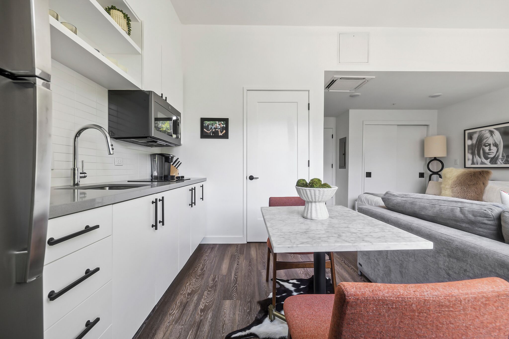 A modern kitchen with white cabinets, stainless steel appliances, and a marble countertop. There is a dining area with a white table and a gray sofa.
