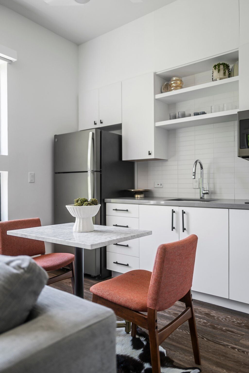 A modern kitchen with white cabinetry, a stainless steel refrigerator, and a built-in oven. There is a dining area with a marble table and two chairs.