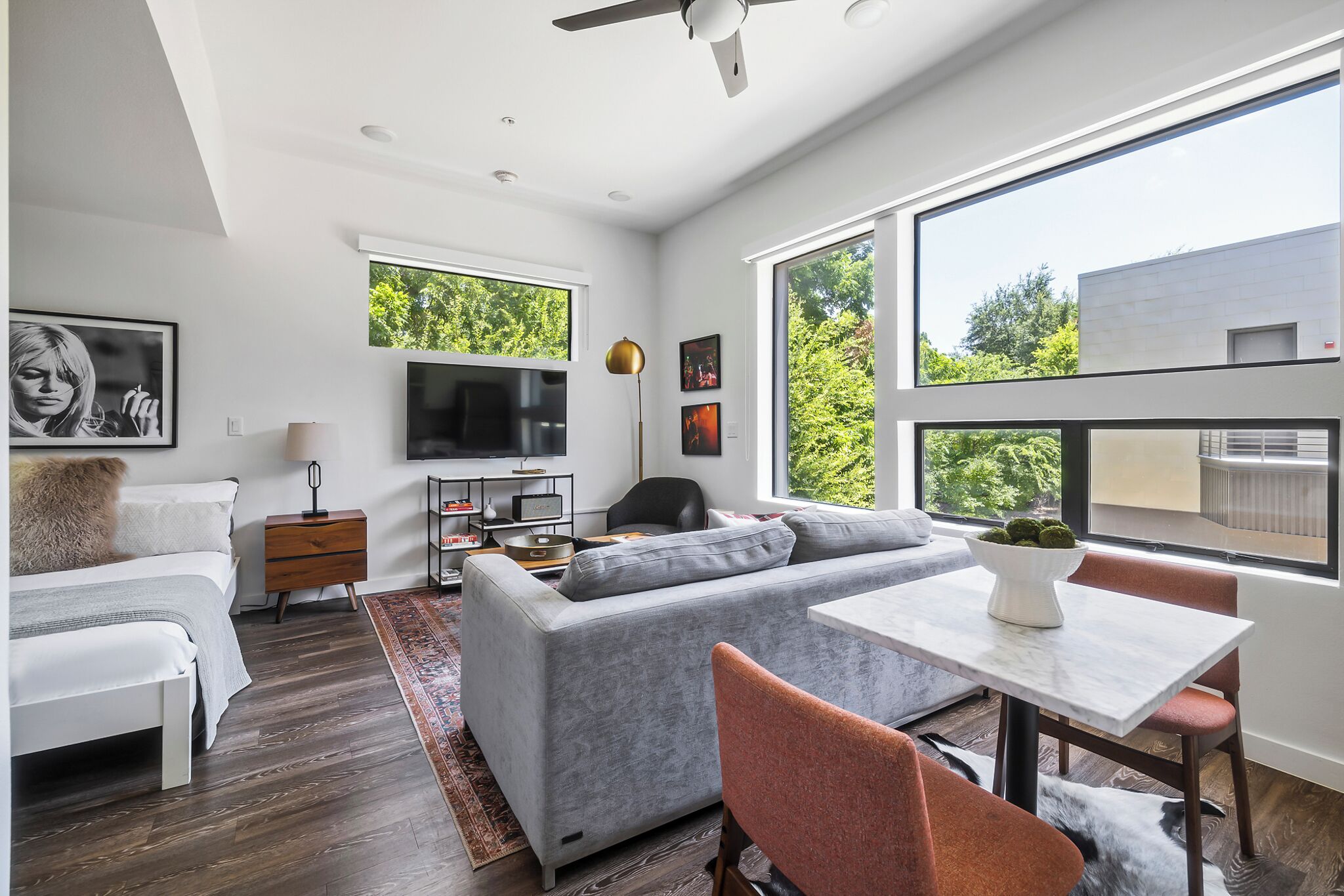 A modern living room with a grey sofa, a white coffee table, and a TV. There is a window with a view of trees and a building.