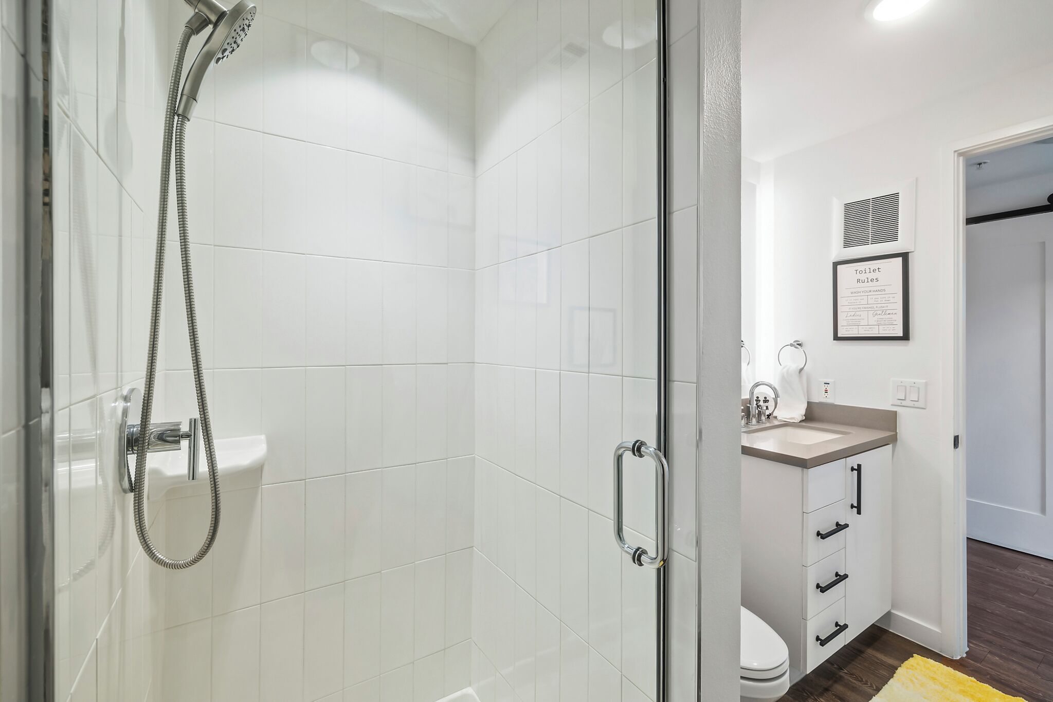 Un baño moderno con cabina de ducha de vidrio, un lavabo blanco y un cartel con normas enmarcado en la pared.