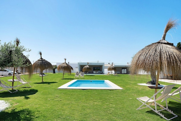 A swimming pool surrounded by grass, with lounge chairs and thatched umbrellas.
