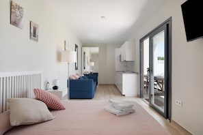 A modern hotel room with a bed, a blue sofa, a small kitchenette, and a sliding glass door to the outside.