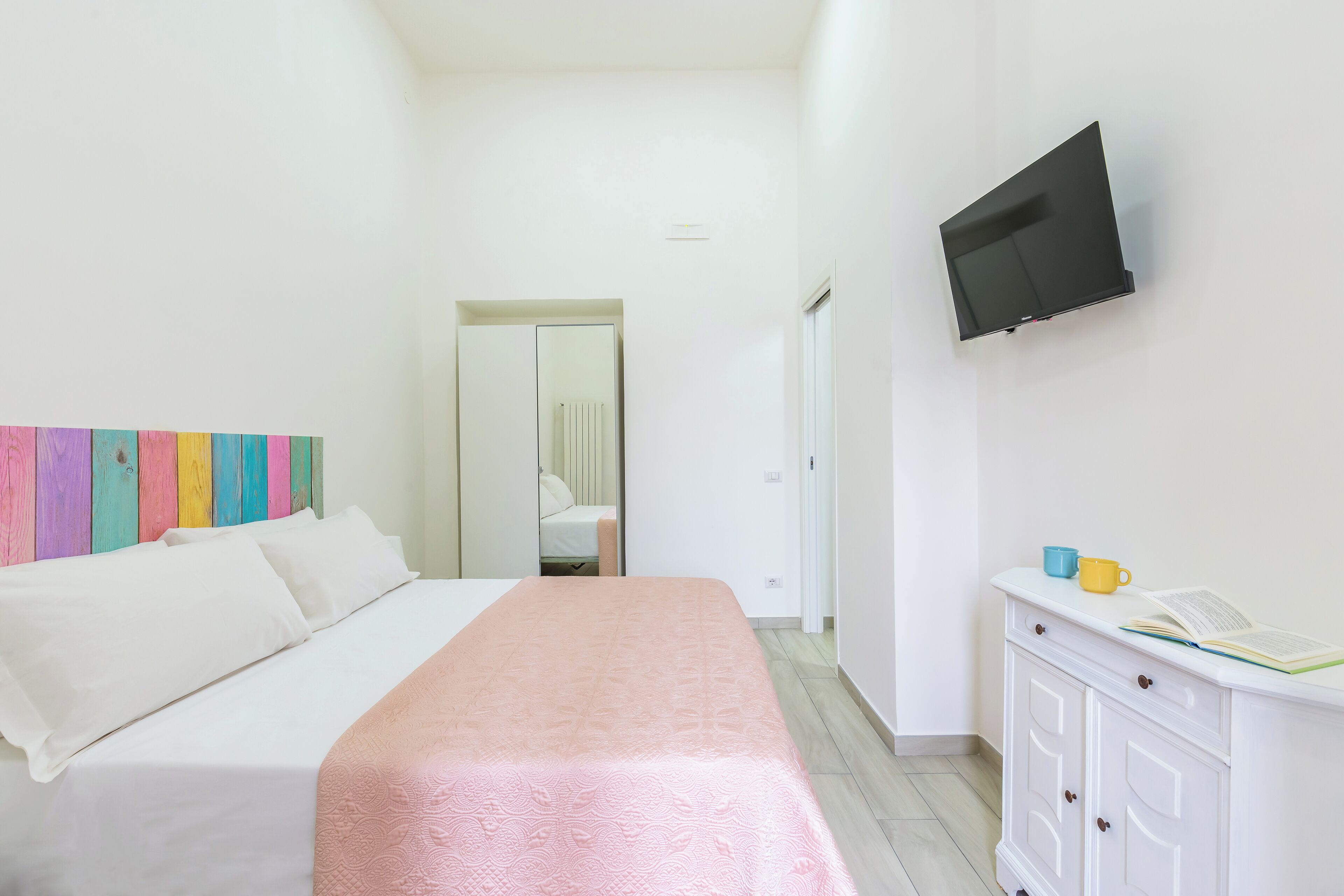 A hotel room with a bed, a TV mounted on the wall, a bedside table with a book and a mug, and a colorful wooden headboard.