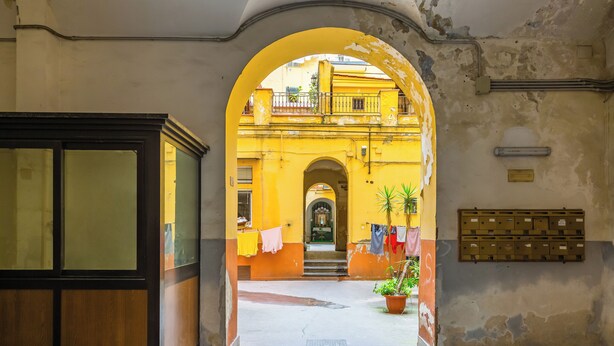 An arched doorway leading to a courtyard with a yellow building, a mailbox, and a potted plant.