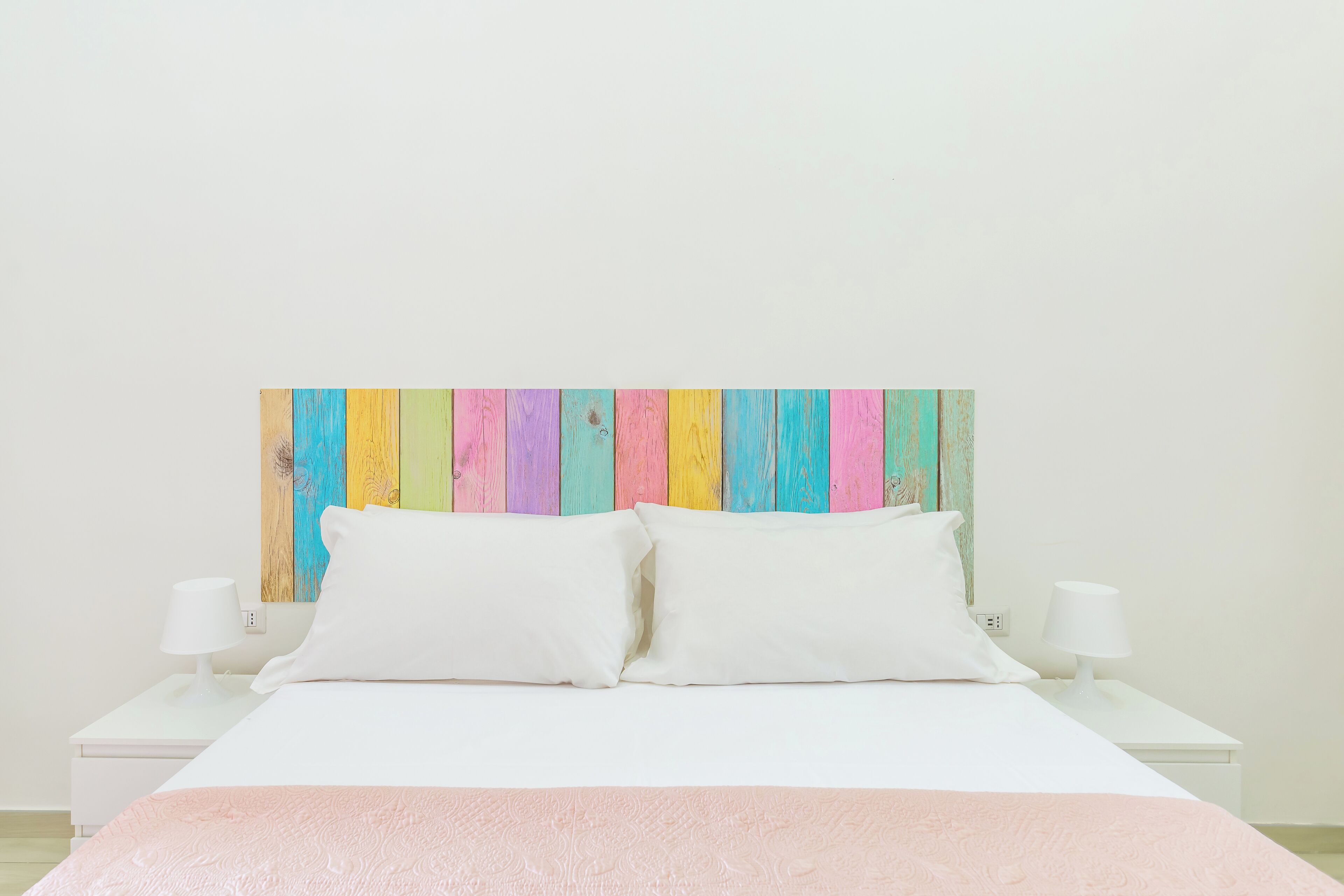Un lit avec une literie blanche et deux oreillers blancs, une tête de lit en bois aux couleurs vives et des tables de chevet blanches avec des lampes.