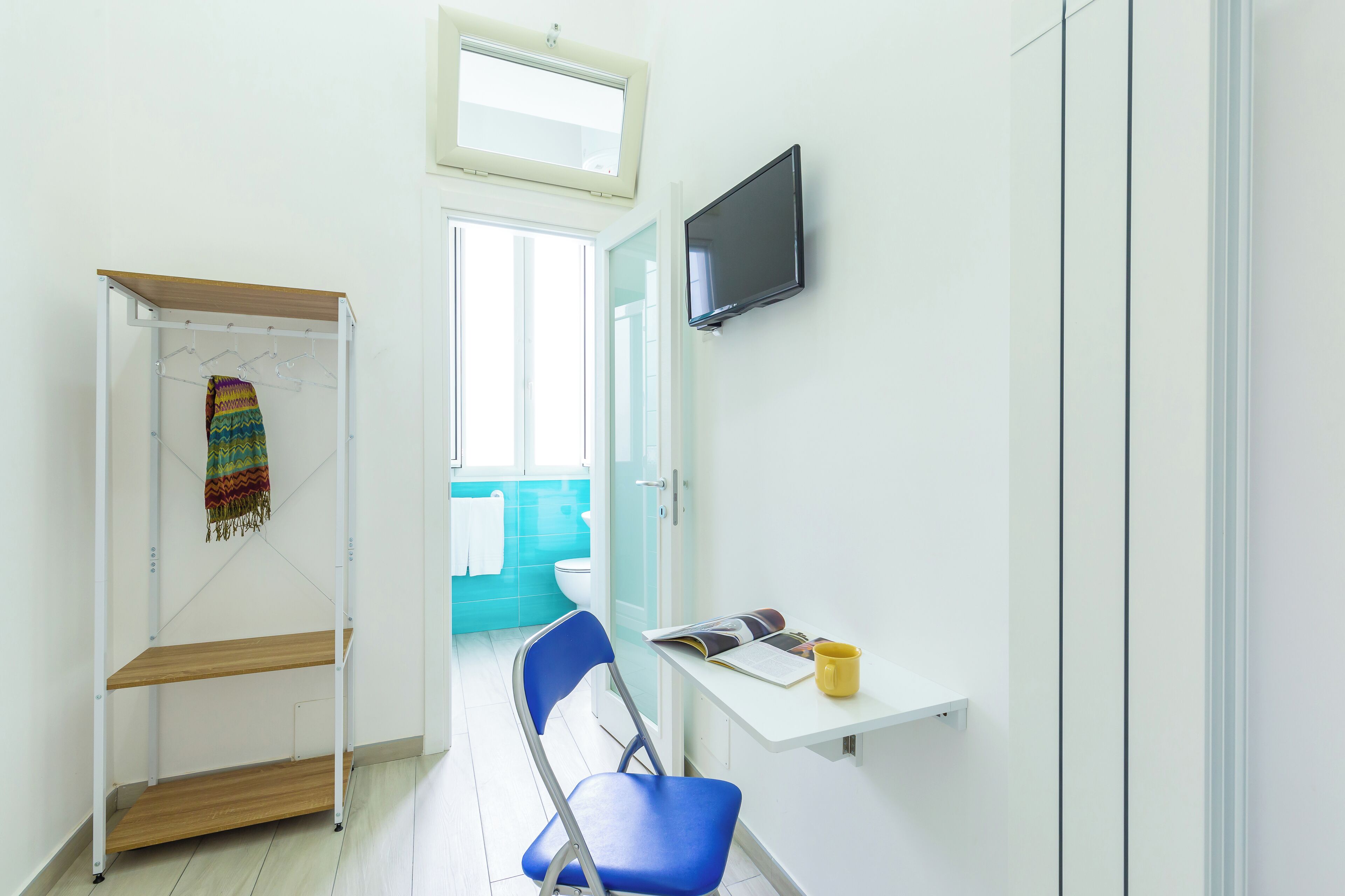 A small, bright room with a white desk, a blue chair, a shelf with a colorful towel, and a TV on the wall.