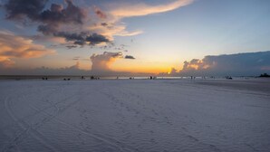 Une vaste plage ouverte, parsemée d’empreintes de pas qui mènent vers l’eau, et un coucher de soleil en arrière-plan.
