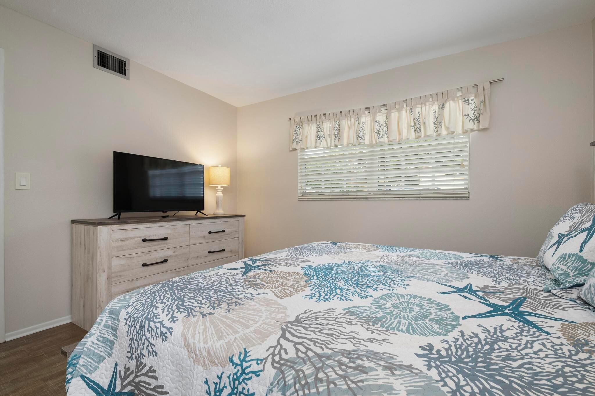 A bedroom with a bed, a TV on a wooden dresser, a window with blinds, and a wall vent.