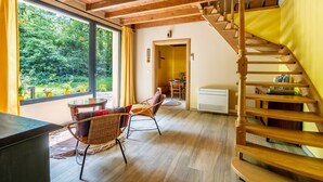 A room with a wooden staircase, a dining area, and a view of greenery outside.