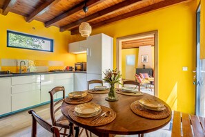 A dining area with a wooden table set for four, surrounded by chairs, a vase with flowers, and a kitchen in the background. - Forest Cottage 'Maison forestière Pleine Nature Kerdary' with Private Terrace, Garden & Wi-Fi (Nivillac)