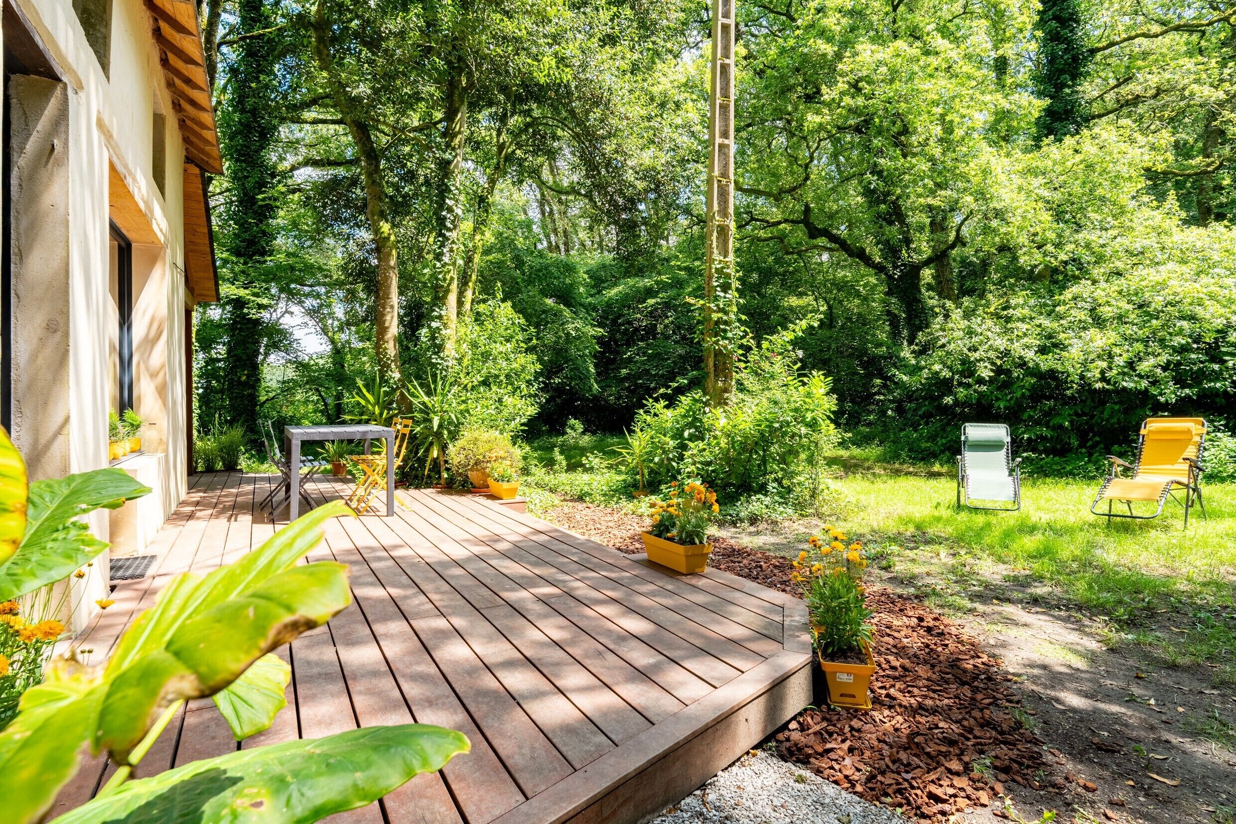 A wooden deck with potted plants, a table, and chairs, surrounded by lush greenery and trees.