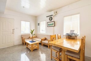Une pièce comprenant une table à manger en bois, des chaises, un canapé et une petite table sur laquelle est posée une plante. On y trouve également des fenêtres avec des stores, une porte et une horloge fixée au mur.