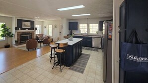 Een ruime woonkamer met een open haard, een eethoek met een groot kookeiland, en een keuken met donkere kastjes en een koelkast.